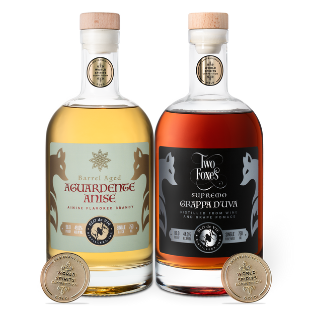Two bottles of aged aguardente anise and grappa d'uva with labels and San Francisco World Spirits Competition gold medals on a white background